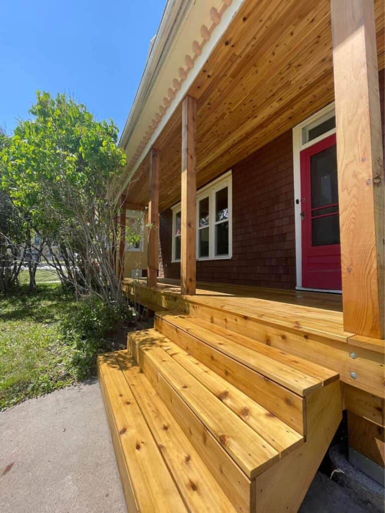 Front Porch Decking Front porch with wooden steps and railing, attached to a brick house with a red door.