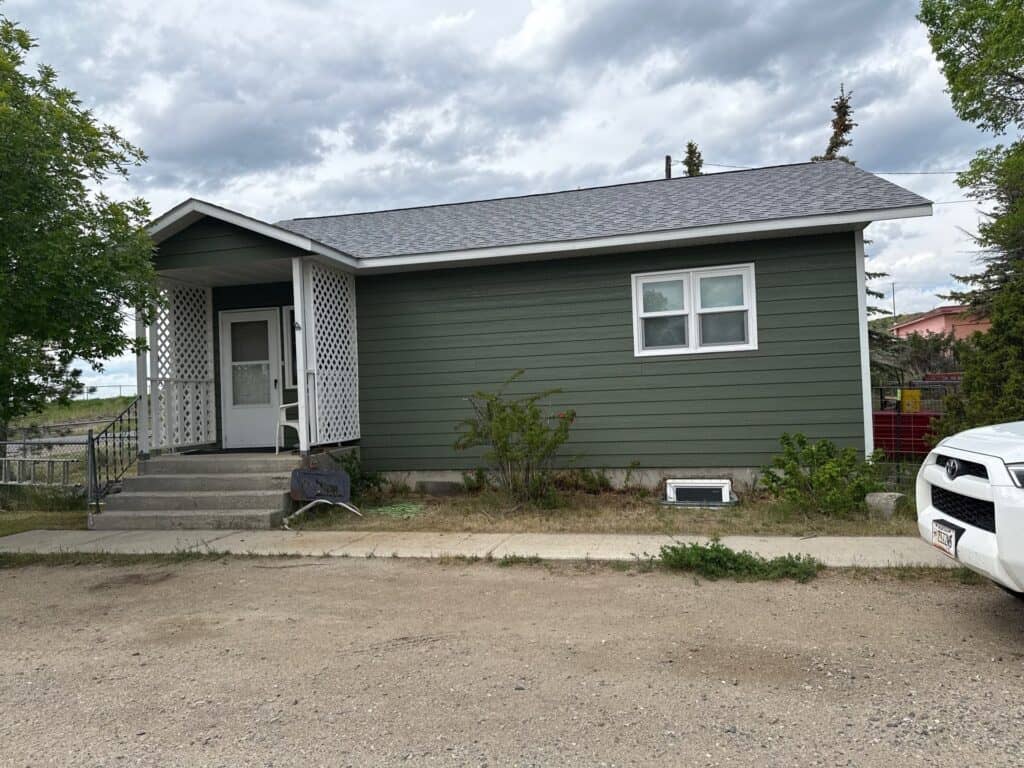 Single-story green house with front porch and white window frames, located in a residential area.
