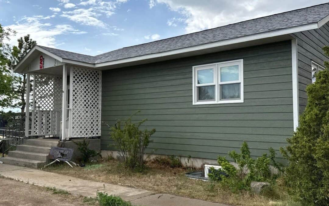 Single-story green house with white trim and front porch, surrounded by bushes and a concrete walkwa.