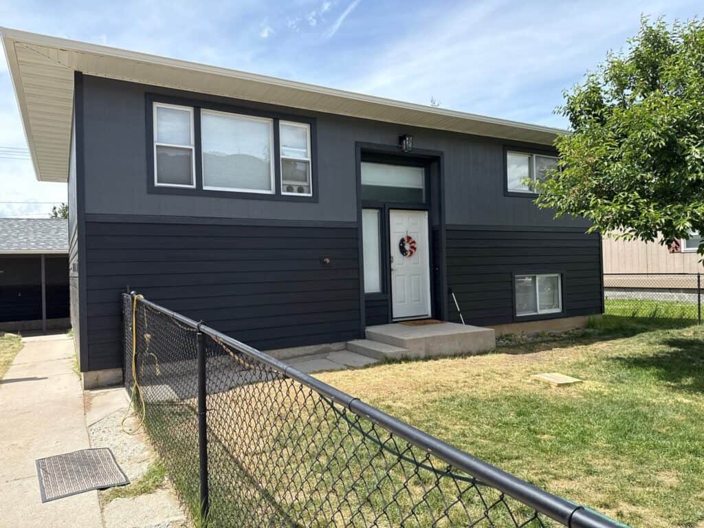 Contemporary house with dark siding and large windows, front yard with grass and tree.