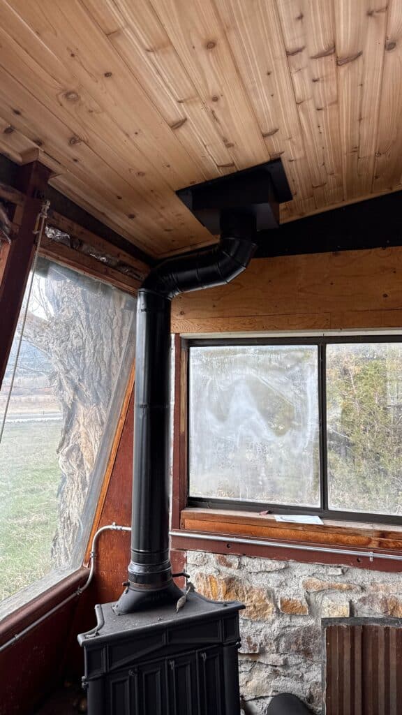 Wood stove with chimney pipe in cozy indoor space with large window and stone wall.