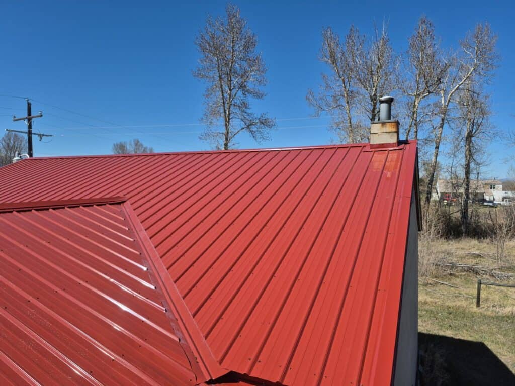 Red metal roof on a building with a chimney and clear blue sky.
