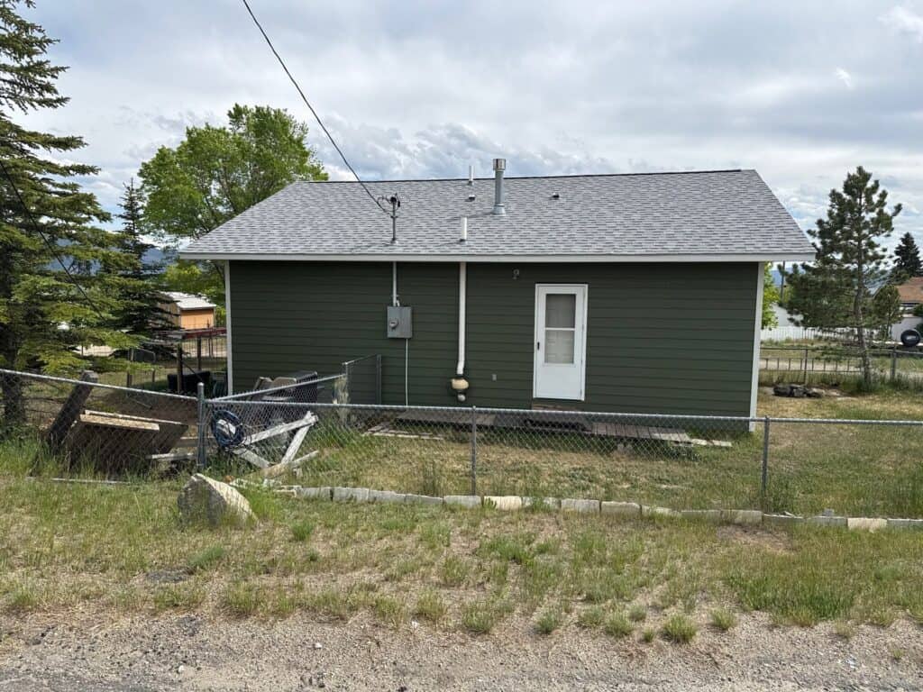 Green house with gray roof and white door in a yard with trees and fence.